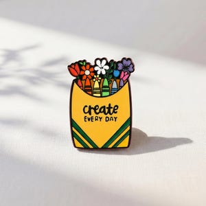 May include: A yellow enamel pin shaped like a crayon box with green diagonal stripes at the bottom. The box contains colorful flowers and crayons, with the black text create EVERY DAY printed in the center of the yellow surface.