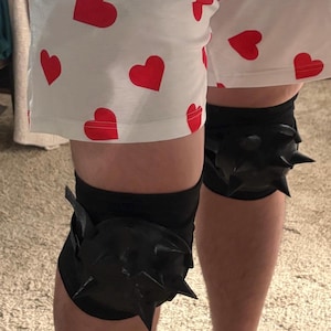 May include: White shorts featuring a repeating pattern of small red hearts, paired with black knee pads that have prominent, sharp, black spikes protruding from the center of each pad, worn over the knees.