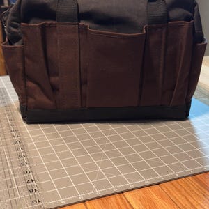 May include: A black and brown tote bag with multiple pockets. The bag has a rectangular shape with a black base and handles. The front features several brown pockets. The bag is sitting on a gray cutting mat.