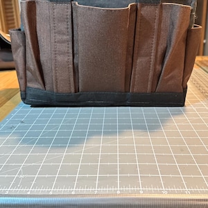 Può includere: Una borsa porta attrezzi in tela marrone e nera con più tasche e un manico. La borsa ha una forma rettangolare ed è progettata per trasportare attrezzi e forniture. La borsa è appoggiata su un tappetino da taglio grigio.