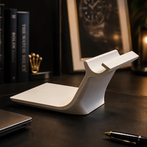 May include: A white, modern phone or tablet stand with a curved base and a raised ledge to hold a device. The stand is made of a solid, smooth material and sits on a dark surface. A laptop and pen are also visible.