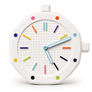 May include: A white wall clock with an octagonal frame and a textured grid dial. The clock features colorful hour markers in shades of pink, orange, yellow, green, blue, and purple, with light blue hour and minute hands and a white side knob.