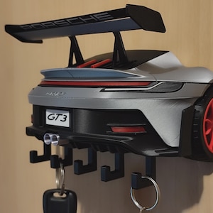 Può includere: Un portachiavi da parete a forma di parte posteriore di una Porsche 911 GT3 color argento. Il modello presenta un alettone nero con la scritta PORSCHE, ruote rosse, doppio scarico e una targa con la scritta GT3. Cinque ganci neri reggono anelli portachiavi.