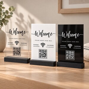 May include: Three rectangular signs with black wooden bases. Each sign features the text Welcome, Please enjoy your wifi, Network, Password, and a QR code. The first sign is clear acrylic, the second is opaque white, and the third is opaque black.
