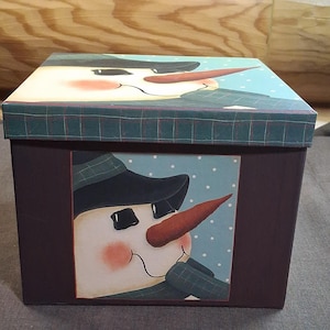 May include: A square storage box with a dark brown base and a lid featuring a green plaid pattern. The front and top panels display a painted illustration of a snowman with a black hat, a carrot nose, and rosy cheeks against a light blue background.