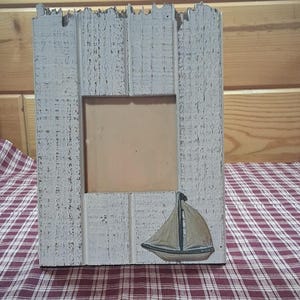 May include: A rectangular picture frame constructed from weathered white wood with a distressed finish. The frame features a square central opening and a painted illustration of a sailboat with tan sails and a dark hull positioned in the lower right corner.