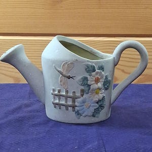 May include: A light gray ceramic watering can planter featuring a raised relief design of a dragonfly, a wooden-style fence, and a cluster of white and blue flowers with green leaves. The vessel has a curved handle and a wide, open top opening.