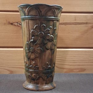 May include: A tall, cylindrical ceramic vase featuring a brown and dark green glazed finish. The exterior is decorated with embossed floral patterns, vertical ribbed detailing, and a scalloped rim, creating a textured appearance throughout.
