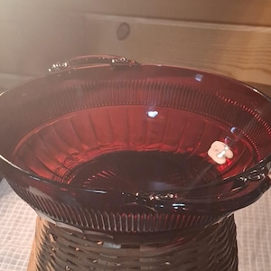 May include: A ruby red glass bowl with a ribbed design and decorative handles. The bowl is set on a woven wicker base. The bowl's color is a deep, rich red, and it appears to be vintage.