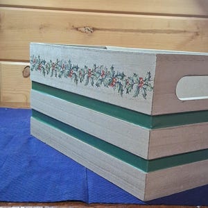 May include: Wooden crate with a light gray finish, featuring a green stripe design and a holly berry garland. The crate has a handle on one side and is placed on a blue surface. Ideal for holiday decor.