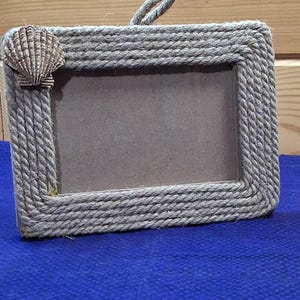 May include: A rectangular picture frame wrapped in thick, natural-colored jute rope. The frame features a single, textured, fan-shaped seashell attached to the upper left corner. A small rope loop is attached to the top center for hanging purposes.