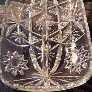 May include: Clear glass divided serving dish with starburst and geometric patterns. The rectangular dish has a scalloped edge and is divided into two sections. The dish is likely vintage and sits on a dark blue patterned surface.