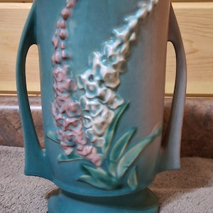 May include: A teal-colored ceramic vase with two handles and a pedestal base. The vase features raised floral designs in pink and white, with green leaves. The vase has a slightly distressed appearance.