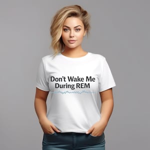 May include: White t-shirt with the text "Don't Wake Me During REM" in black, above a blue waveform design. The shirt is a crew neck style.