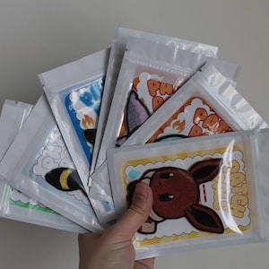 May include: A hand holds a fan of several sealed plastic pouches. Each pouch contains a colorful patch with a cartoon character design. The patches feature various colors and designs, including a brown and white character with the word "CATCH".