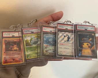 Op de afbeelding: Een hand die verschillende beoordeelde Pokémon-kaarten in beschermende hoesjes vasthoudt. De kaarten tonen verschillende Pokémon-personages, waaronder Charizard, Bulbasaur en Pikachu. Elke kaart zit in een transparante plastic houder met een zilveren sluiting.
