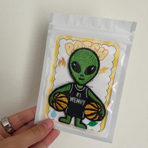 Könnte beinhalten: Ein gestickter Aufnäher mit einer grünen Alien-Figur, die ein schwarzes Basketballtrikot mit dem Text WEMBY und der Nummer 1 trägt und zwei orangefarbene Basketbälle hält. Der Aufnäher befindet sich auf einer gelb-weißen Karte in einem Beutel.