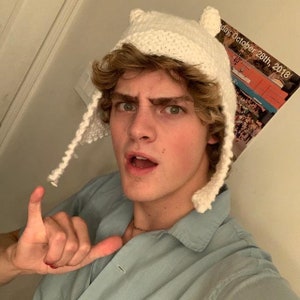 May include: A young person with blond hair wearing a white knitted hat with ear flaps. The hat has a pointed top and resembles an animal's head. The person is wearing a light blue shirt and is making a hand gesture.