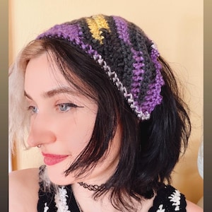 May include: A handmade crochet headband in shades of purple, gray, and yellow. The headband is worn on the head. The crochet work has a textured appearance, with a white and gray border. The overall style is bohemian and casual.