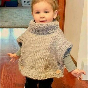 May include: A young child wearing a gray knitted poncho with wooden buttons. The poncho has a turtleneck collar and is made with a chunky yarn.