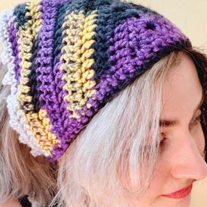 May include: A handmade crochet beanie in a striped pattern of purple, yellow, gray, and white. The beanie is worn on the head and has a textured appearance. The color scheme is vibrant and eye-catching, suitable for fall or winter wear.