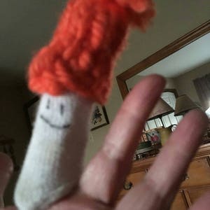 May include: A white finger puppet with a smiling face wearing a bright orange knitted hat.