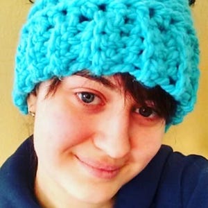 Puede incluir: Una mujer con el pelo oscuro en un moño desordenado lleva una boina de punto de color turquesa brillante. Lleva una sudadera con capucha azul oscuro.