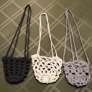 May include: Three handmade crochet bags in a row. The bags are in three different colors: dark gray, off-white, and light gray. Each bag has a long strap for carrying over the shoulder. The bags have an open, net-like design.