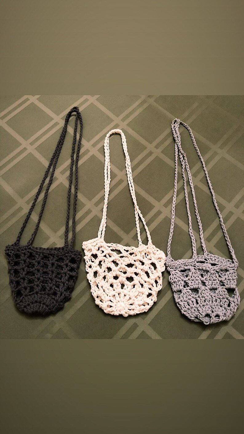 May include: Three handmade crochet bags in a row. The bags are in three different colors: dark gray, off-white, and light gray. Each bag has a long strap for carrying over the shoulder. The bags have an open, net-like design.