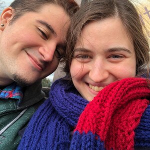 May include: Two people are smiling at the camera. The person on the right is wearing a blue and red knitted scarf.