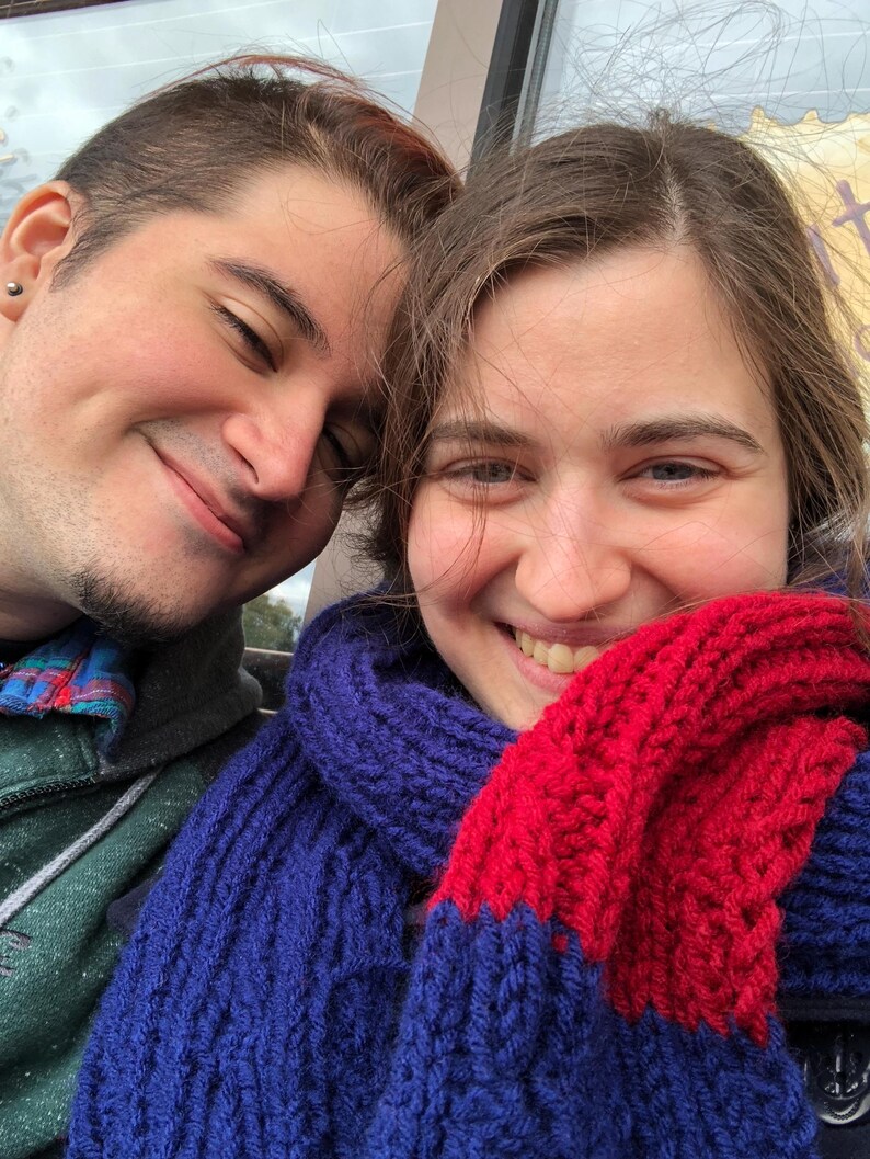 May include: Two people are smiling at the camera. The person on the right is wearing a blue and red knitted scarf.