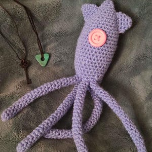 Pu&ograve; includere: Un polpo viola lavorato a uncinetto con un bottone rosa per l'occhio. Il polpo &egrave; sdraiato su una superficie marrone e pelosa. Due collane con una chiave e un ciondolo triangolare turchese sono visibili sullo sfondo. Il testo "Because @knitkritters is the sweetest and most darling friend. My early birthday gift. All Coraline themed" &egrave; visibile nella parte superiore dell'immagine.