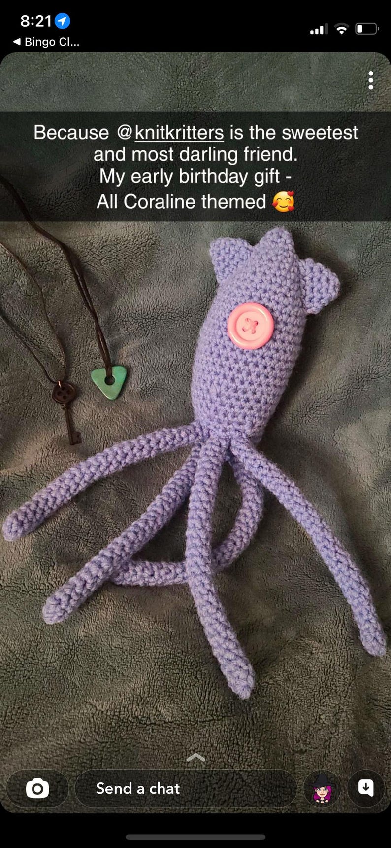 Pu&ograve; includere: Un polpo viola lavorato a uncinetto con un bottone rosa per l'occhio. Il polpo &egrave; sdraiato su una superficie marrone e pelosa. Due collane con una chiave e un ciondolo triangolare turchese sono visibili sullo sfondo. Il testo "Because @knitkritters is the sweetest and most darling friend. My early birthday gift. All Coraline themed" &egrave; visibile nella parte superiore dell'immagine.