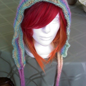 Peut inclure: Une &eacute;charpe &agrave; capuche en tricot color&eacute;e avec de longues attaches. L'&eacute;charpe est faite de plusieurs couleurs, dont le rose, le bleu, le vert et le jaune. L'&eacute;charpe est port&eacute;e par un mannequin aux cheveux longs et rouges.