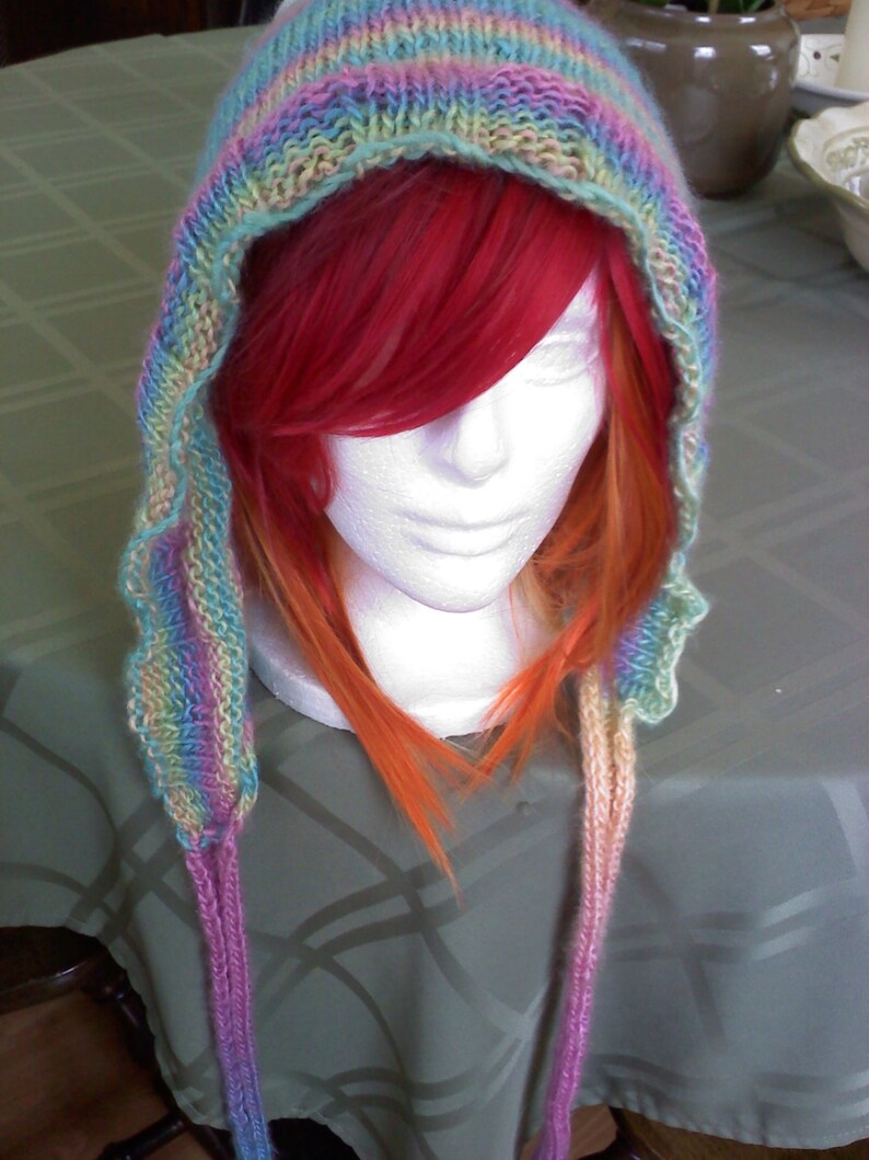 Peut inclure: Une &eacute;charpe &agrave; capuche en tricot color&eacute;e avec de longues attaches. L'&eacute;charpe est faite de plusieurs couleurs, dont le rose, le bleu, le vert et le jaune. L'&eacute;charpe est port&eacute;e par un mannequin aux cheveux longs et rouges.