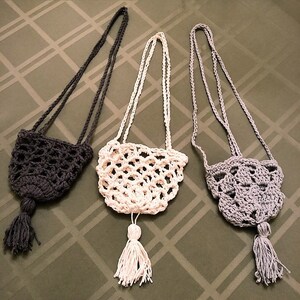 May include: Three handmade crochet bags in dark gray, white, and light gray. Each bag has a long strap and a tassel. The bags are displayed on a patterned green surface.