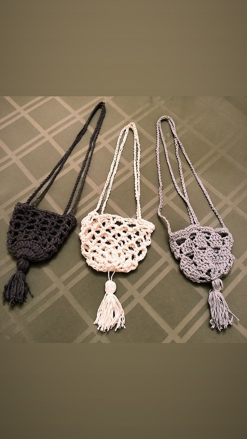 May include: Three handmade crochet bags in dark gray, white, and light gray. Each bag has a long strap and a tassel. The bags are displayed on a patterned green surface.