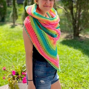 May include: A rainbow crochet shawl with a colorful striped pattern. The shawl is worn by a person with dark hair and is draped around their neck.