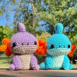 May include: Two crocheted axolotl plushies, one purple and one blue, with orange gills. The plushies are sitting on a wooden surface.