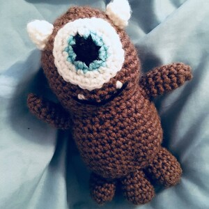 May include: A brown crocheted stuffed toy with a single white eye and a blue and white iris. The toy has two small white ears and brown arms and legs.