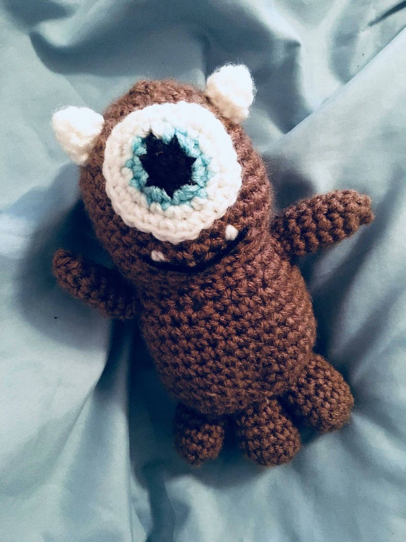 May include: A brown crocheted stuffed toy with a single white eye and a blue and white iris. The toy has two small white ears and brown arms and legs.