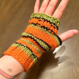 Pode incluir: Luvas sem dedos de malha listrada em laranja, verde e preto. As luvas s&atilde;o feitas com um ponto de tric&ocirc; simples e t&ecirc;m um punho canelado.
