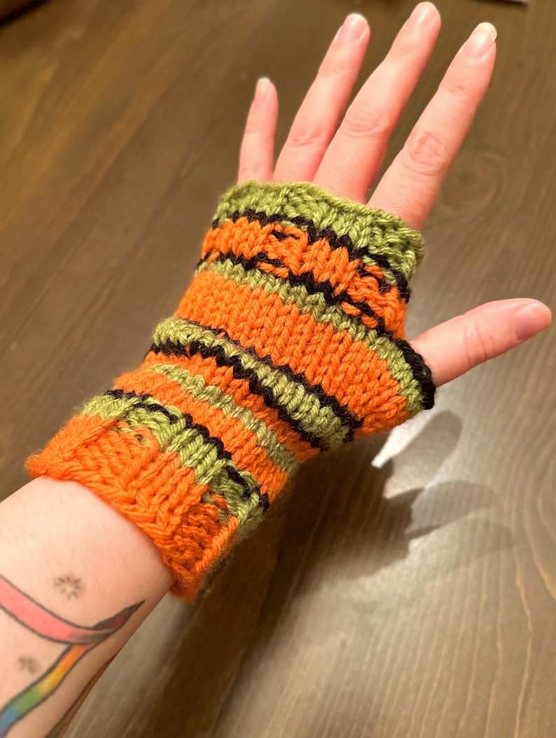 Pode incluir: Luvas sem dedos de malha listrada em laranja, verde e preto. As luvas s&atilde;o feitas com um ponto de tric&ocirc; simples e t&ecirc;m um punho canelado.