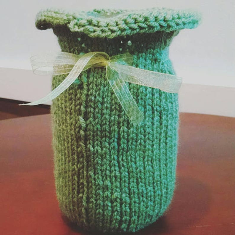 May include: A hand-knitted green cosy with a scalloped top and a sheer ribbon tied around the middle. The cosy is cylindrical and appears to be made of a soft, textured yarn. The ribbon is a pale yellow-green colour.