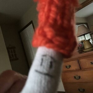 May include: A white sock with a drawn-on face and a bright orange fuzzy hat on top.