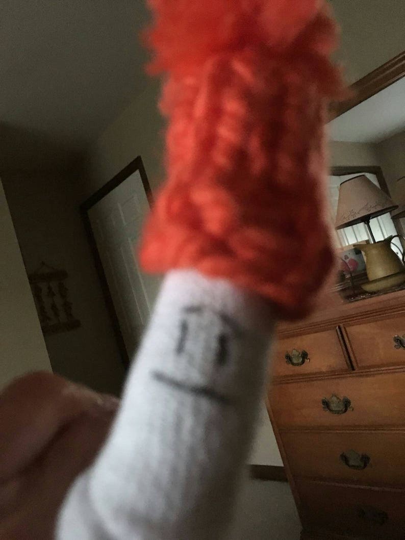 May include: A white sock with a drawn-on face and a bright orange fuzzy hat on top.