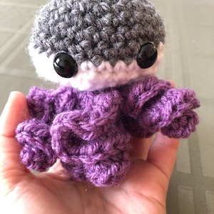 May include: A small crocheted plush toy with a gray head, white face, and purple sweater. The toy has black button eyes.