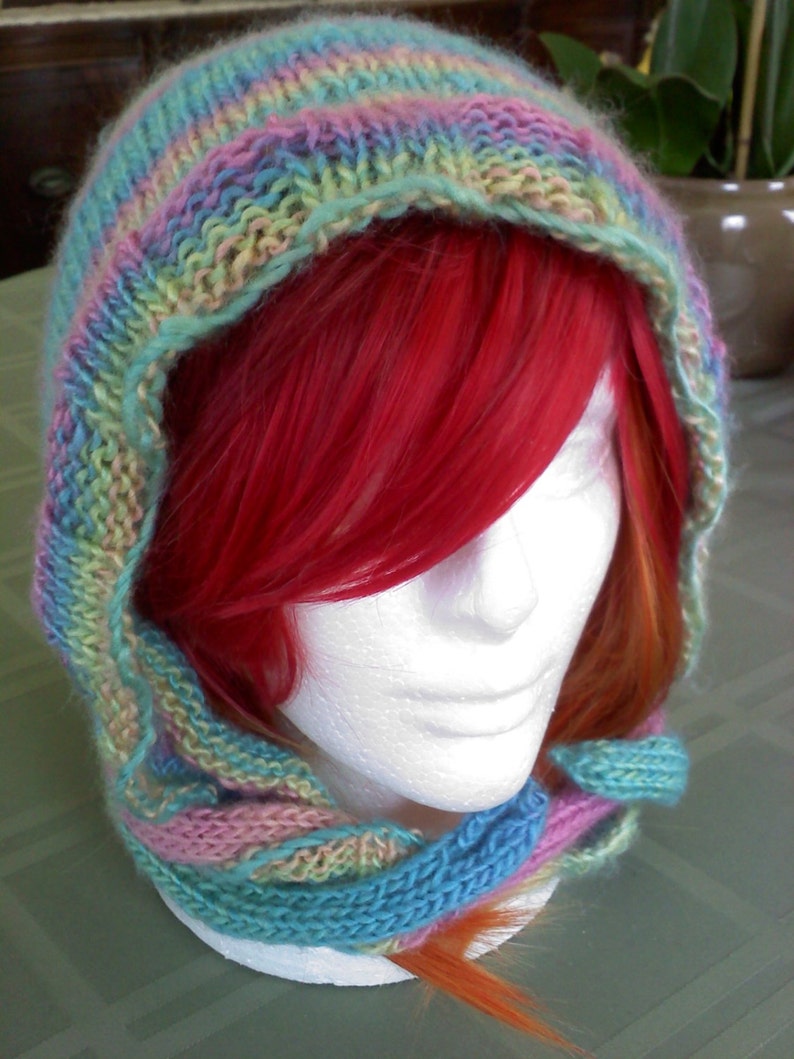 Peut inclure: Une &eacute;charpe &agrave; capuche en tricot dans des tons de rose, bleu et vert. L'&eacute;charpe est port&eacute;e par un mannequin aux cheveux roux et quelques m&egrave;ches orange.