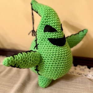 May include: A green crocheted plush toy shaped like a ghost with a stitched-on black mouth and eyes. The toy has a long, thin tail and is standing on a light brown surface.