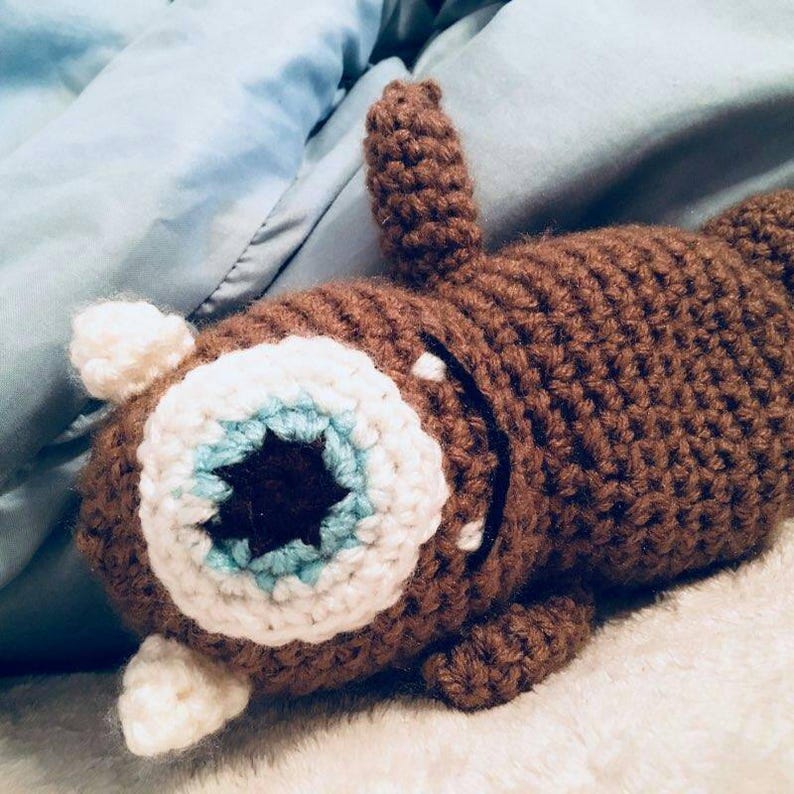 May include: A brown crocheted stuffed toy with a large white eye with a blue and brown centre. The toy is lying on a white surface.